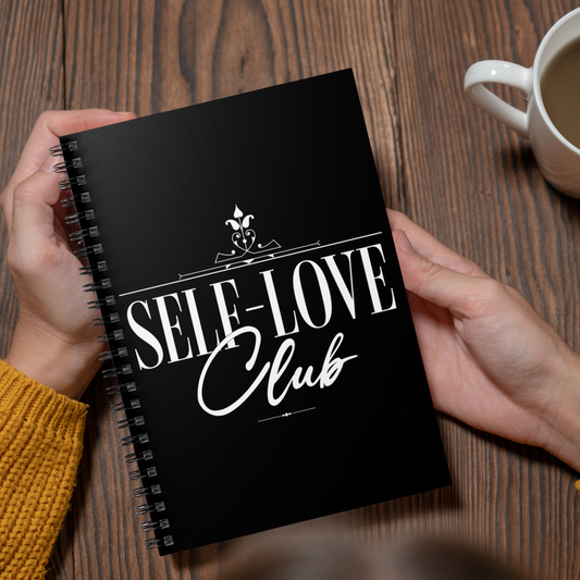 Black notebook titled 'Self-Love Club' held by a person on a wooden table with a cup of coffee.