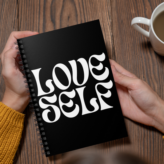 Black notebook with 'LOVE SELF' text held by hands on a wooden table with a cup of coffee.