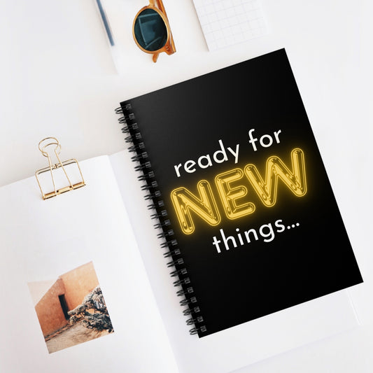 Black notebook with 'ready for NEW things...' text on a white surface with sunglasses and a photo clip.